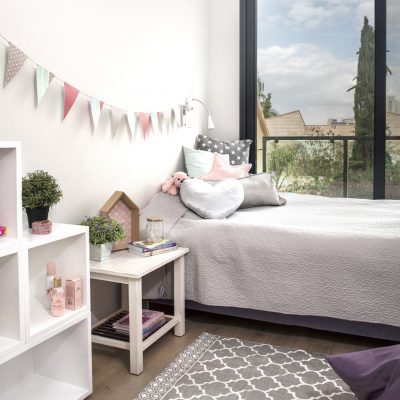 חדר של ילדה עם מיטה בהירה , שטיח אפור ודגלונים צבעוניים על הקיר